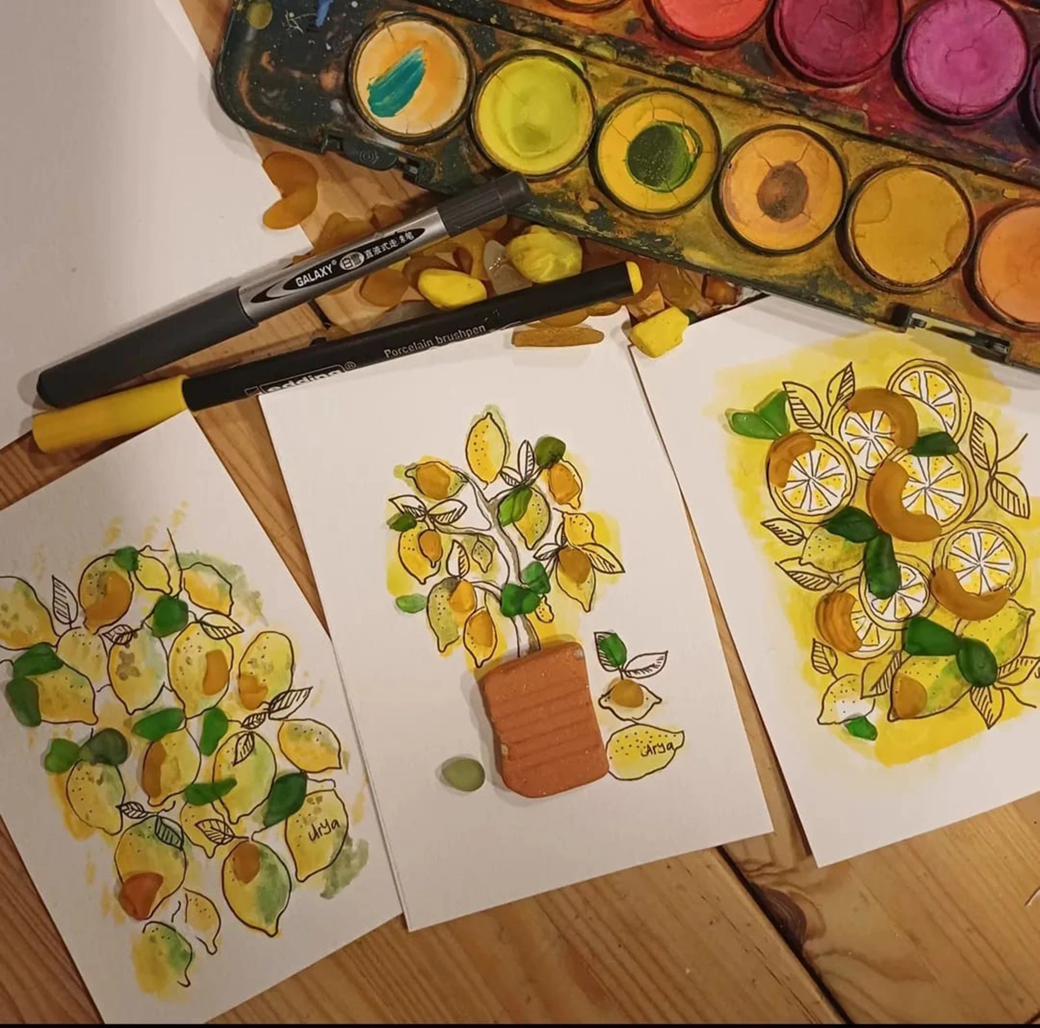 Plano cerrado de la mesa de trabajo de la artista con 3 dibujos de una de sus series terminado y las acuarelas y rotuladores que se han usado para hacer los dibujos