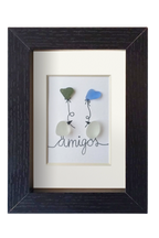 Cuadro de vidrio de mar y tinta en el que dos Pájaros posados encima del texto amigos se intercambian sedos globos con forma de corazón. Marco negro