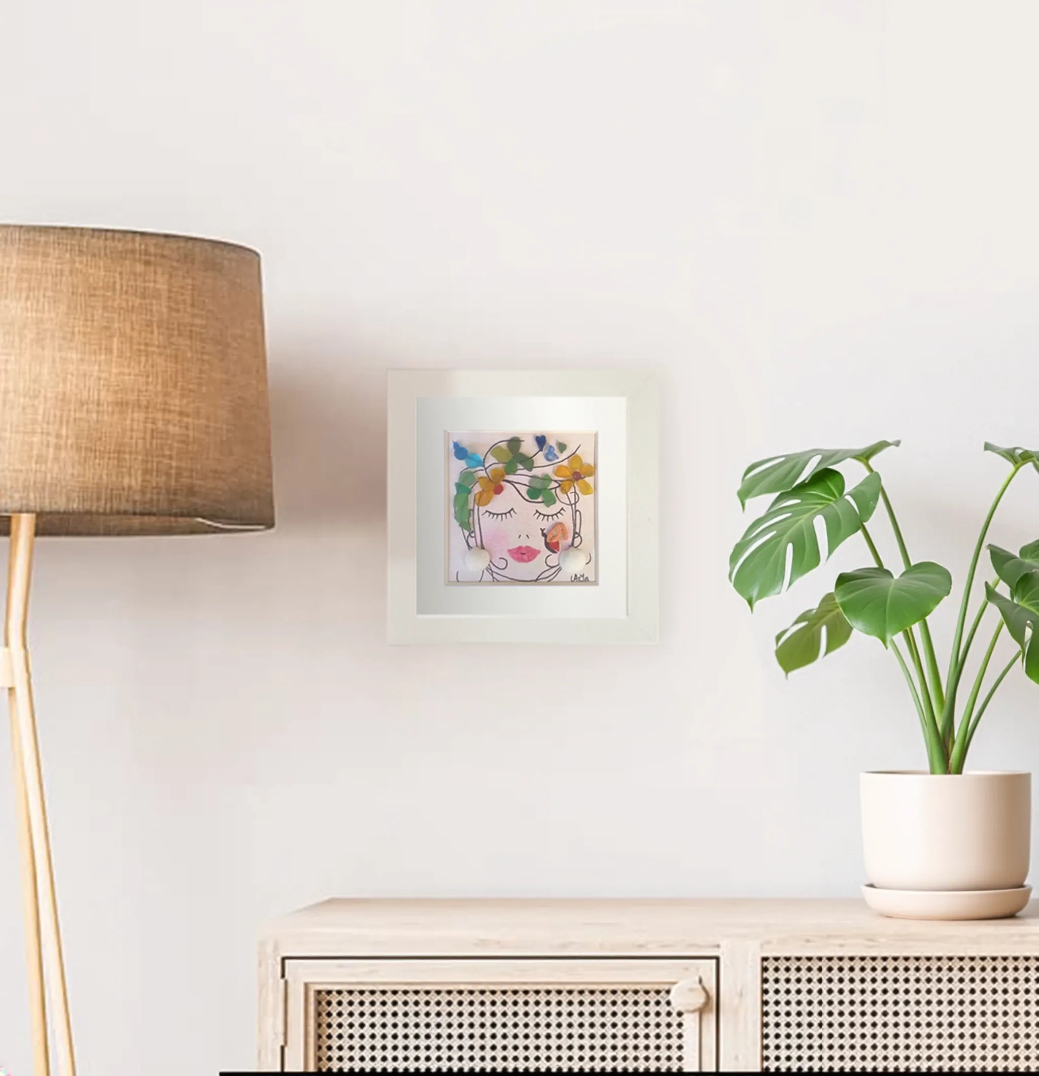 Pared con lámpara de pie saliendo por la izquierda de la imagen, con cuadro de chica soñadora con flores en la cabeza y pendientes de conchas hecha con acuarela y vidrio de mar de colores y mueble bajo de madera y mimbre con una maceta de monstera encima
