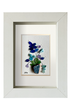 Cuadro de maceta de fragmento de ceramica erosionada de color azul petróleo con una planta de hojas verdes y flores de distintos tonos azules y lila de tinta y vidrios de mar erosionados