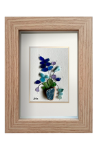 Cuadro de maceta de fragmento de ceramica erosionada de color azul petróleo con una planta de hojas verdes y flores de distintos tonos azules y lila de tinta y vidrios de mar erosionados