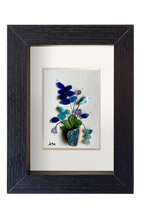 Cuadro de maceta de fragmento de ceramica erosionada de color azul petróleo con una planta de hojas verdes y flores de distintos tonos azules y lila de tinta y vidrios de mar erosionados