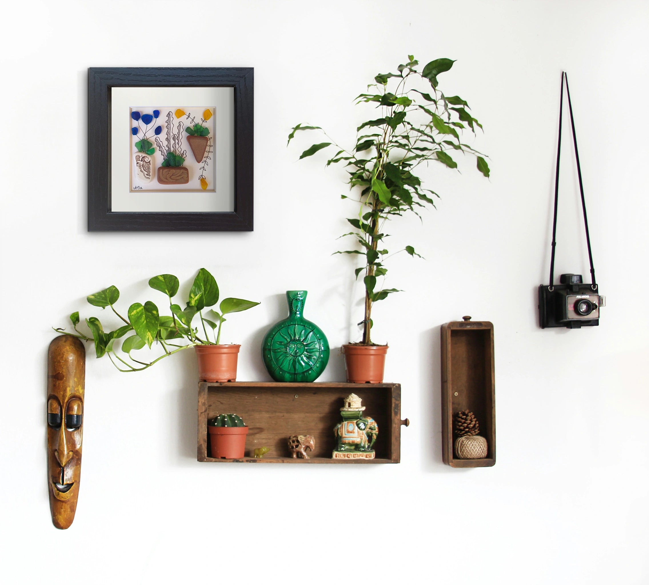 Pared blanca con diversos elementos colgados: 2 cajones antiguos y un cuadro de vidrio de mar con flores, una cámara, una máscara de madera y otros elementos decorativos 