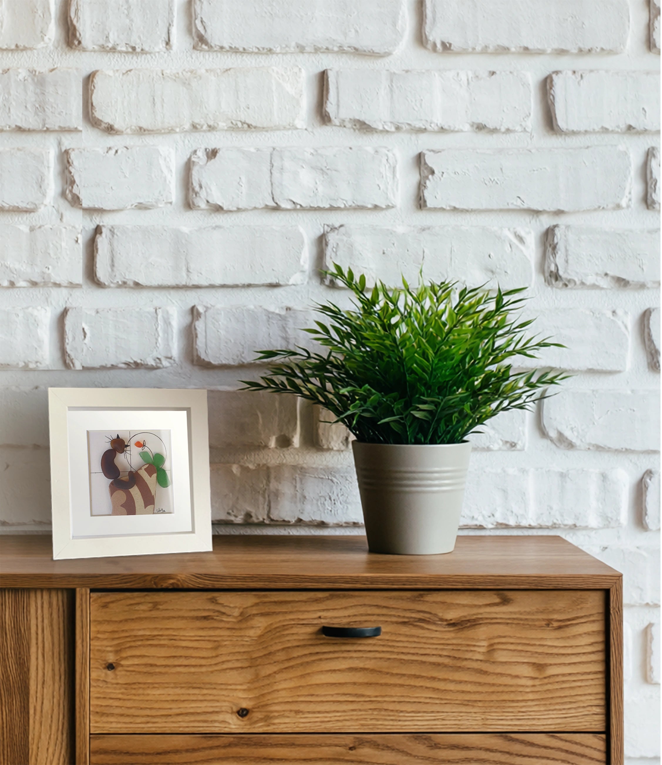 Pared de ladrillos blanca con cómoda de madera con un cuadro de vidrio de mar y una planta encima