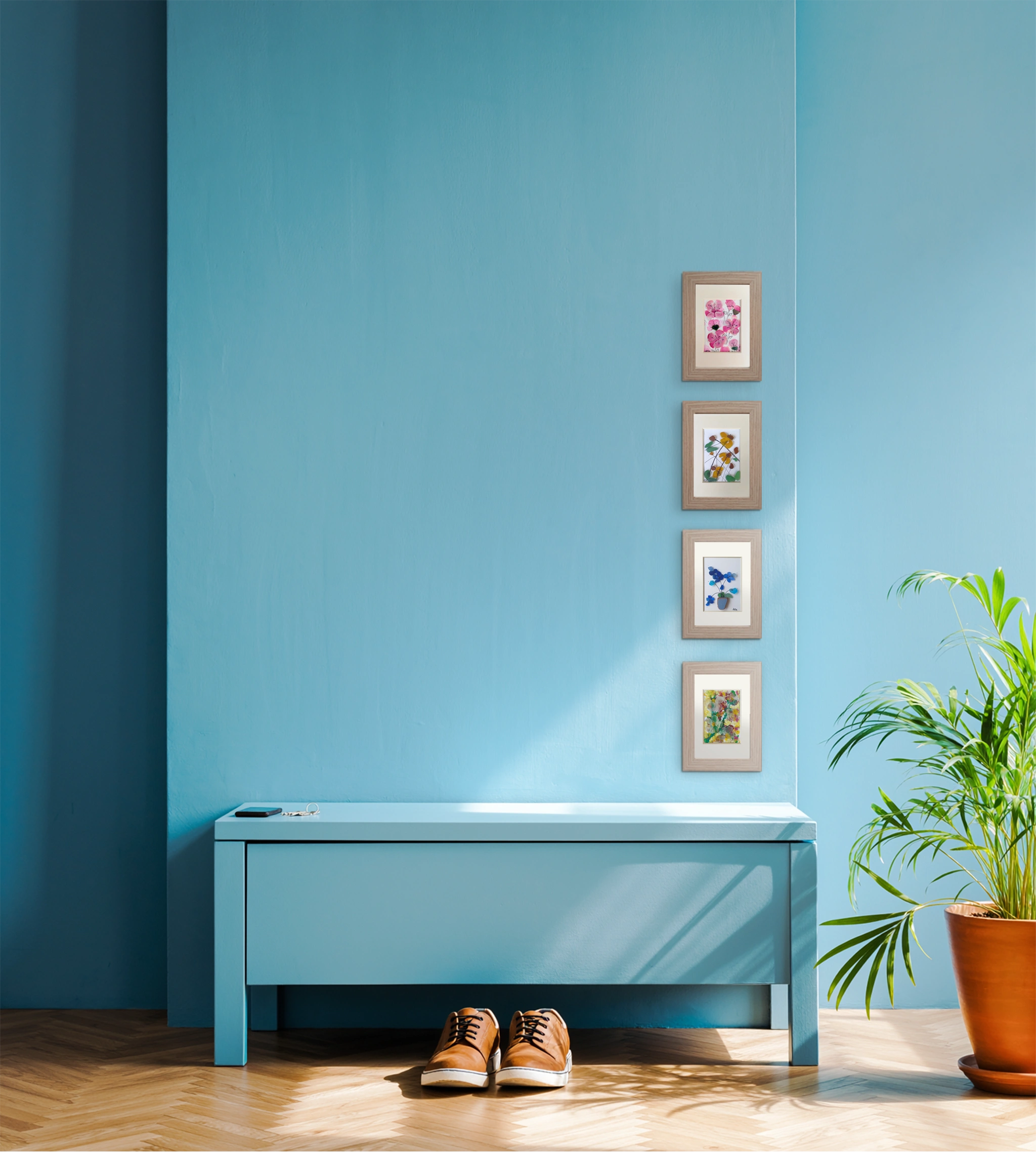 Imagen de pared de recibidor azul celeste con mueble bajo del mismo color, con llaves y unos zapatos en el suelo. Por la derecha aparece una bonita planta. 4 cuadros de vidrio de mar erosionado con representaciones florales cuelgan la pared decorándola