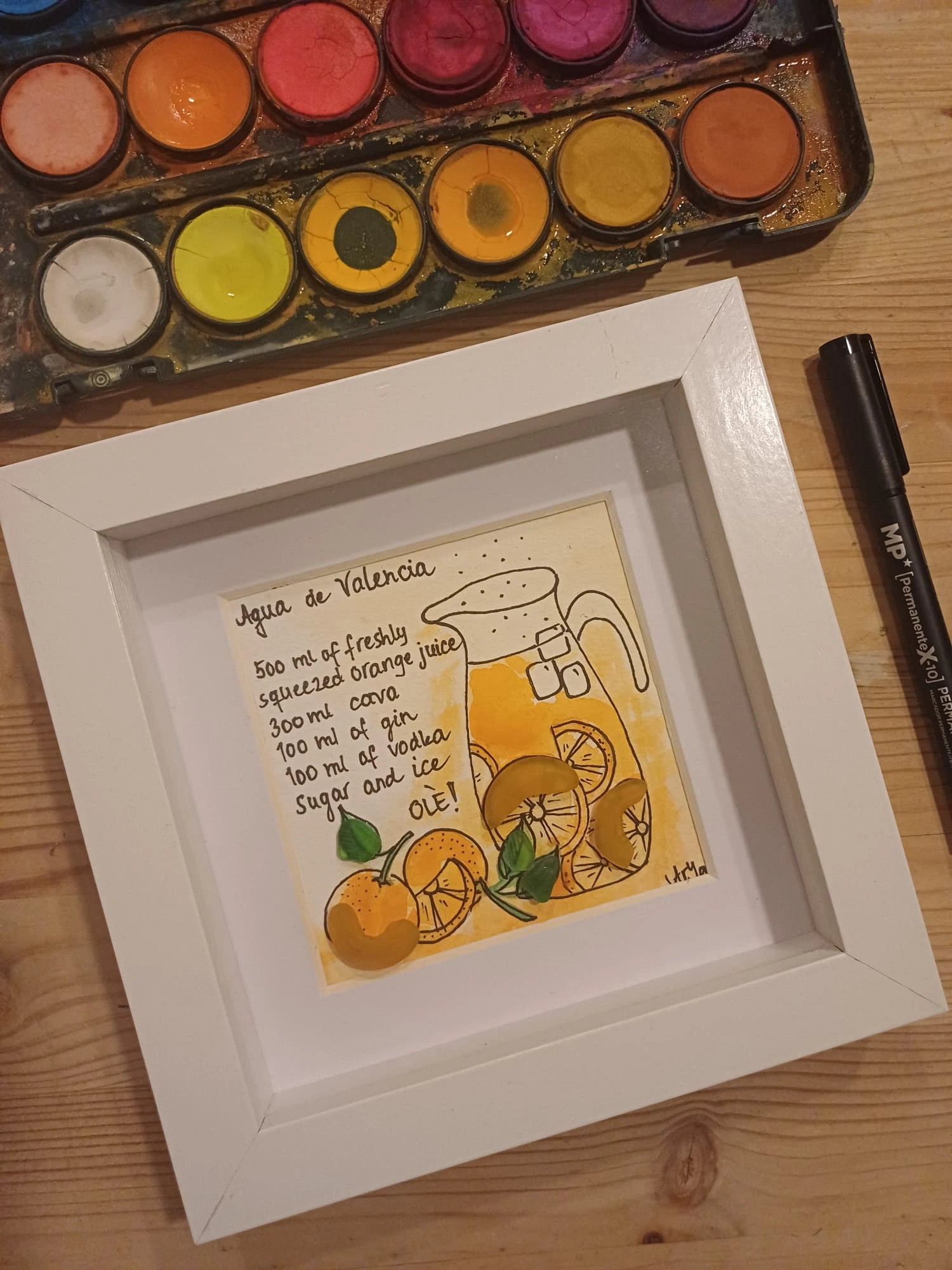 Plano cerrado de la mesa de trabajo de la artista con uno de sus cuadros terminado y las acuarelas y el rotulador que se han usado para hacer el cuadro