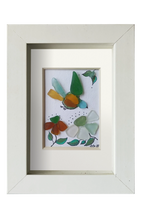 Cuadro de vidrio de mar y tinta con un pajaro multicolor volando hacia unas flores. Marco blanco